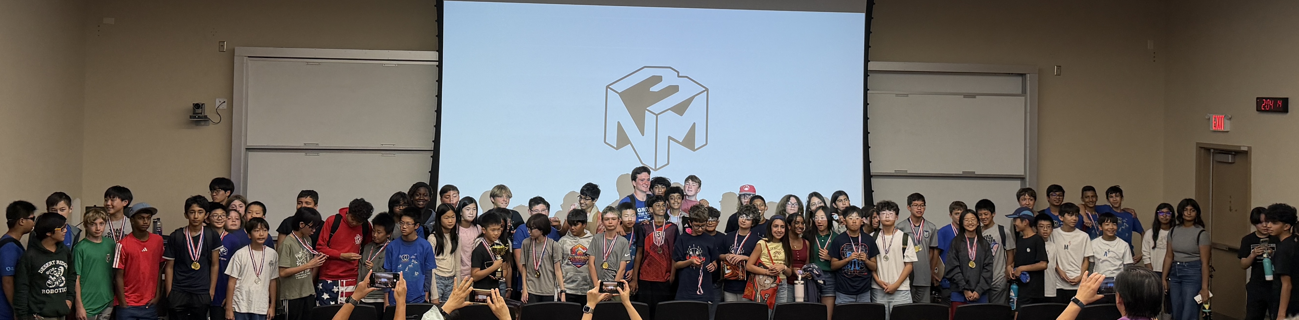 About 100 middle school students, many wearing medals, pose together in front of a screen showing the NM3 cube logo as parents take photos.