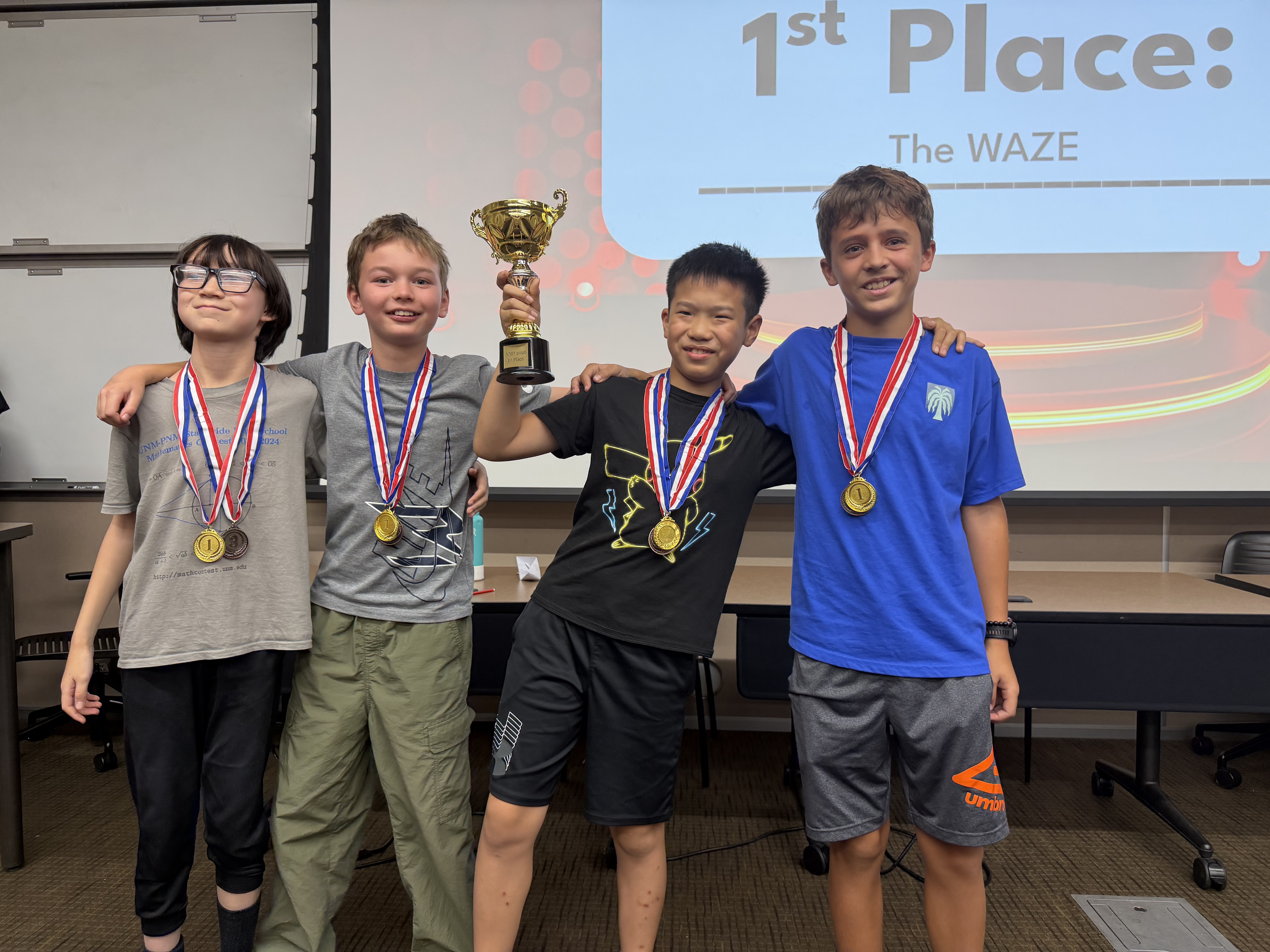 Four smiling students wearing gold medals stand arm in arm before a screen reading 1st Place: The WAZE; one raises a gold trophy.