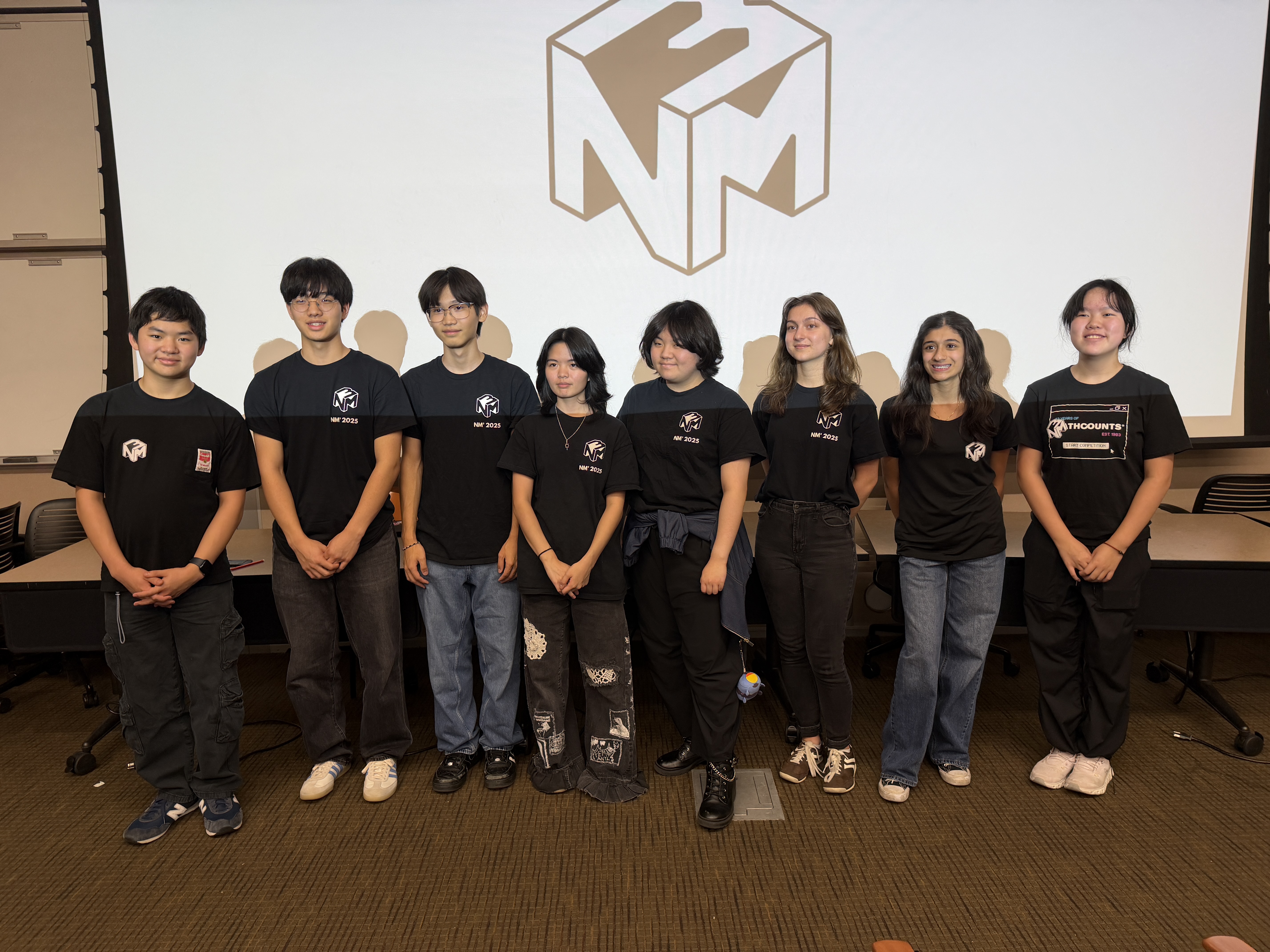 Eight high school students in matching black NM3 T-shirts stand in a row in front of a screen showing the NM3 cube logo.